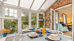 A conservatory with a dining table set for a meal, surrounded by large windows and a brick wall. - 3 Bed in Docking (oc-g29874) (Docking)