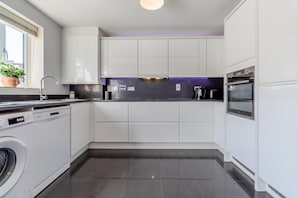 A modern kitchen with white cabinets, a dishwasher, a washing machine, a built-in oven, and a window with a plant.