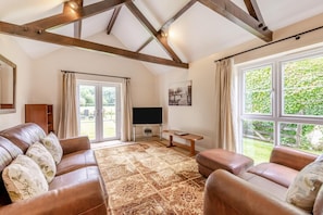 A spacious living room with a large leather sofa, a coffee table, and a flat-screen TV. There is a large window providing a view of the outdoors. - 3 Bed in Two Waters Foot (oc-p29856) (Broadoak)