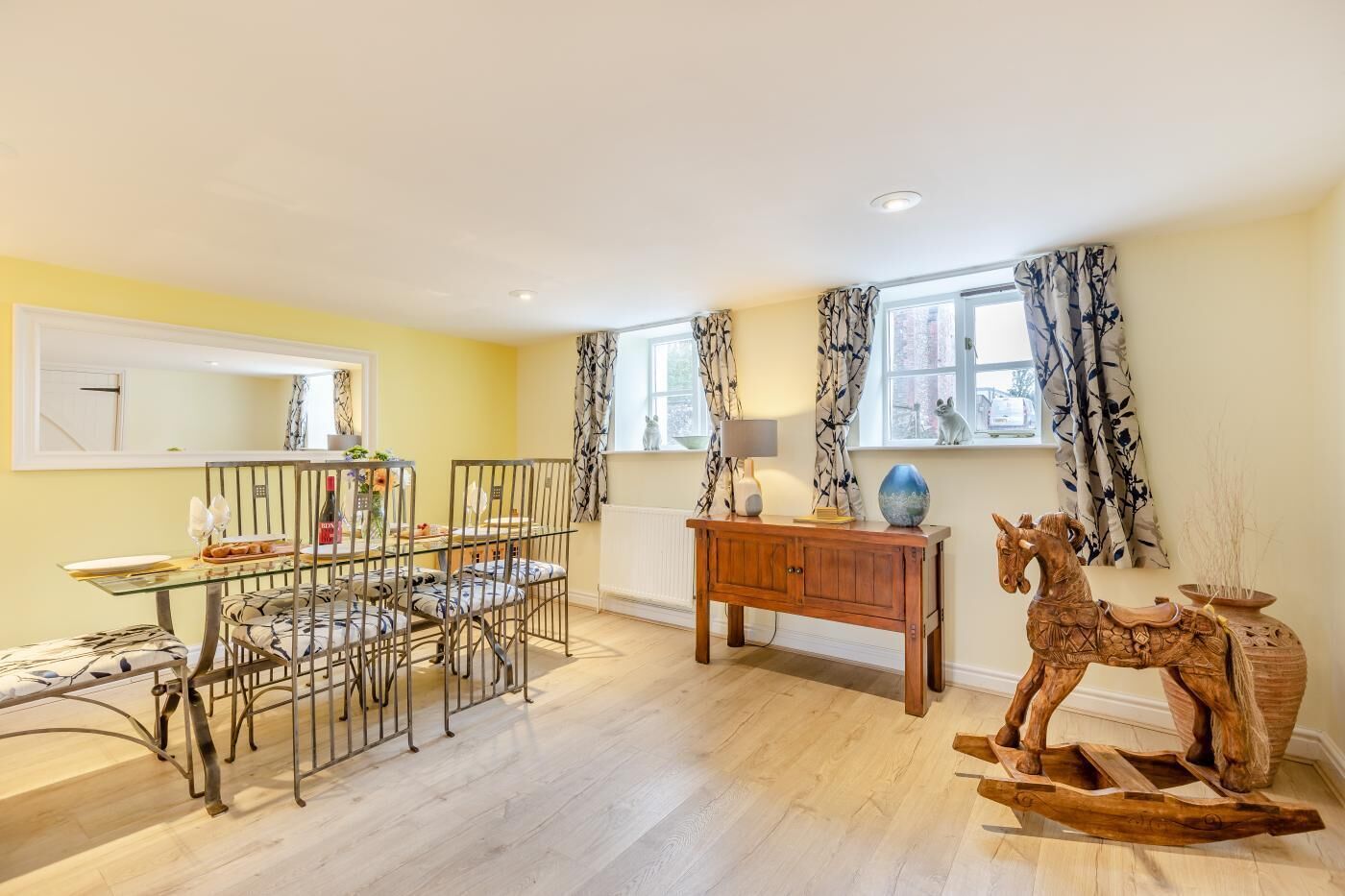 A dining area with a glass table and metal chairs, a wooden side table with a lamp, and a decorative horse sculpture.