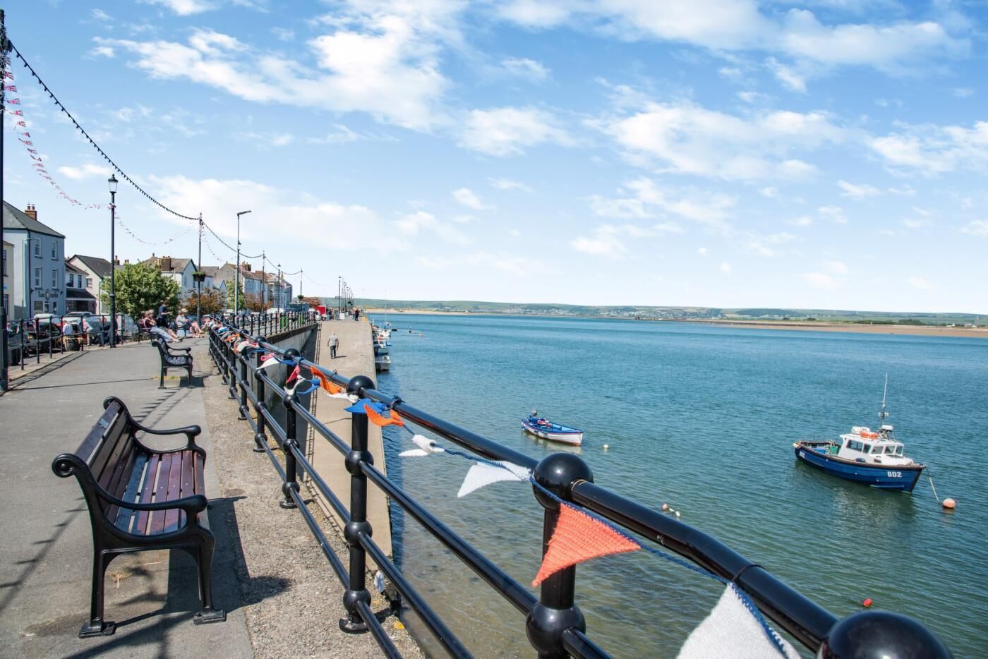 Une promenade en bord de mer, agrémentée de bancs, d’une rambarde et de bateaux sur l’eau.