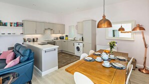 Une cuisine moderne avec des meubles blancs, un coin repas doté d’une table en bois pour quatre personnes, et un canapé bleu agrémenté de coussins roses.