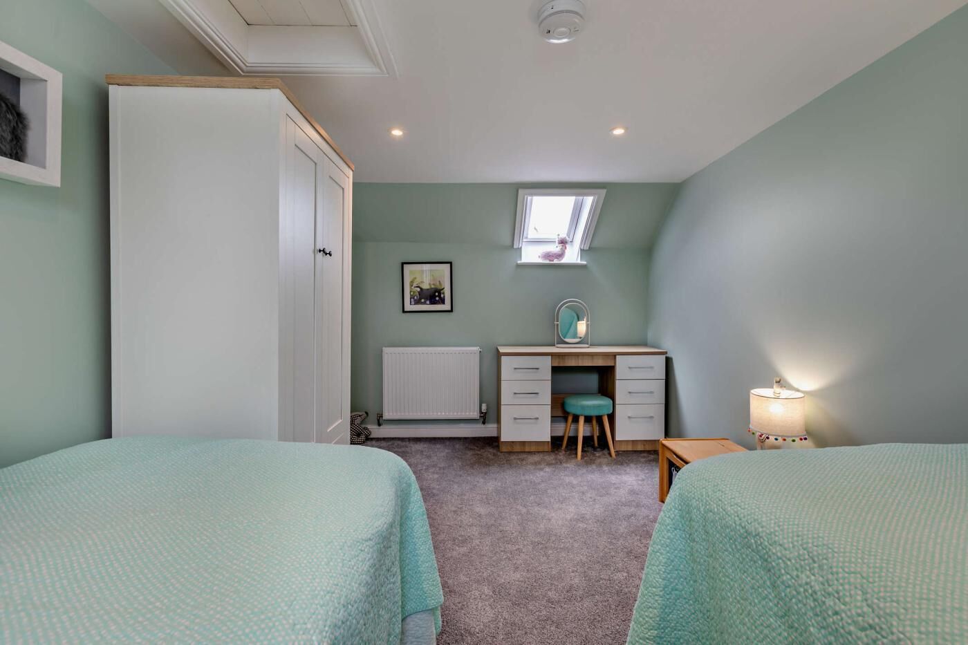 A bedroom with two beds, a desk with a chair, a radiator, and a window.