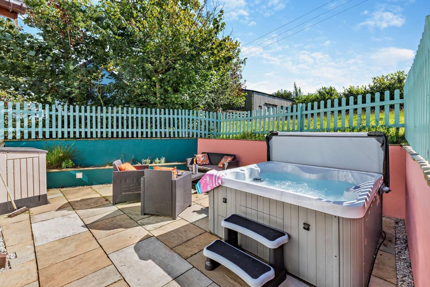 A hot tub with steps, wicker furniture, and a teal fence in a backyard setting.