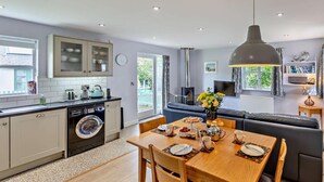 Une cuisine moderne équipée d’un lave-linge, un coin repas avec une table dressée pour deux personnes, et un salon avec un canapé et un téléviseur.