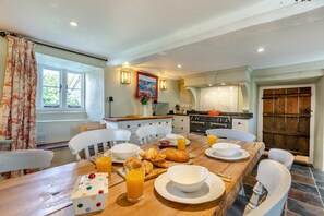 Uma sala de jantar com uma mesa de madeira, cadeiras brancas, itens de café da manhã e uma cozinha ao fundo.