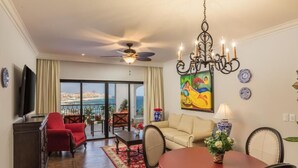 Un salón amplio con un sillón rojo, sofá beige y una mesa de comedor con sillas. Un candelabro cuelga del techo, y hay un cuadro en la pared.