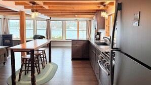 A modern kitchen with wooden beams, large windows, a dining area, and a view of a lake.