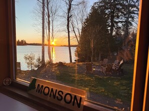 A window with a view of a lake at sunset, a deck with chairs, and a sign reading 'MONSON'.