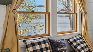 A room with two windows overlooking a lake, beige curtains, and checkered pillows on a bench.