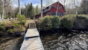 A wooden dock leading to a house with a red exterior, surrounded by trees and a body of water.
