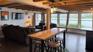 A cabin interior with a wooden dining table, bar stools, a sofa, a TV mounted on the wall, and large windows overlooking a lake.
