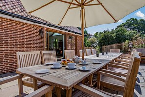 An outdoor dining area with a long wooden table and chairs, a large umbrella, and a brick building in the background.