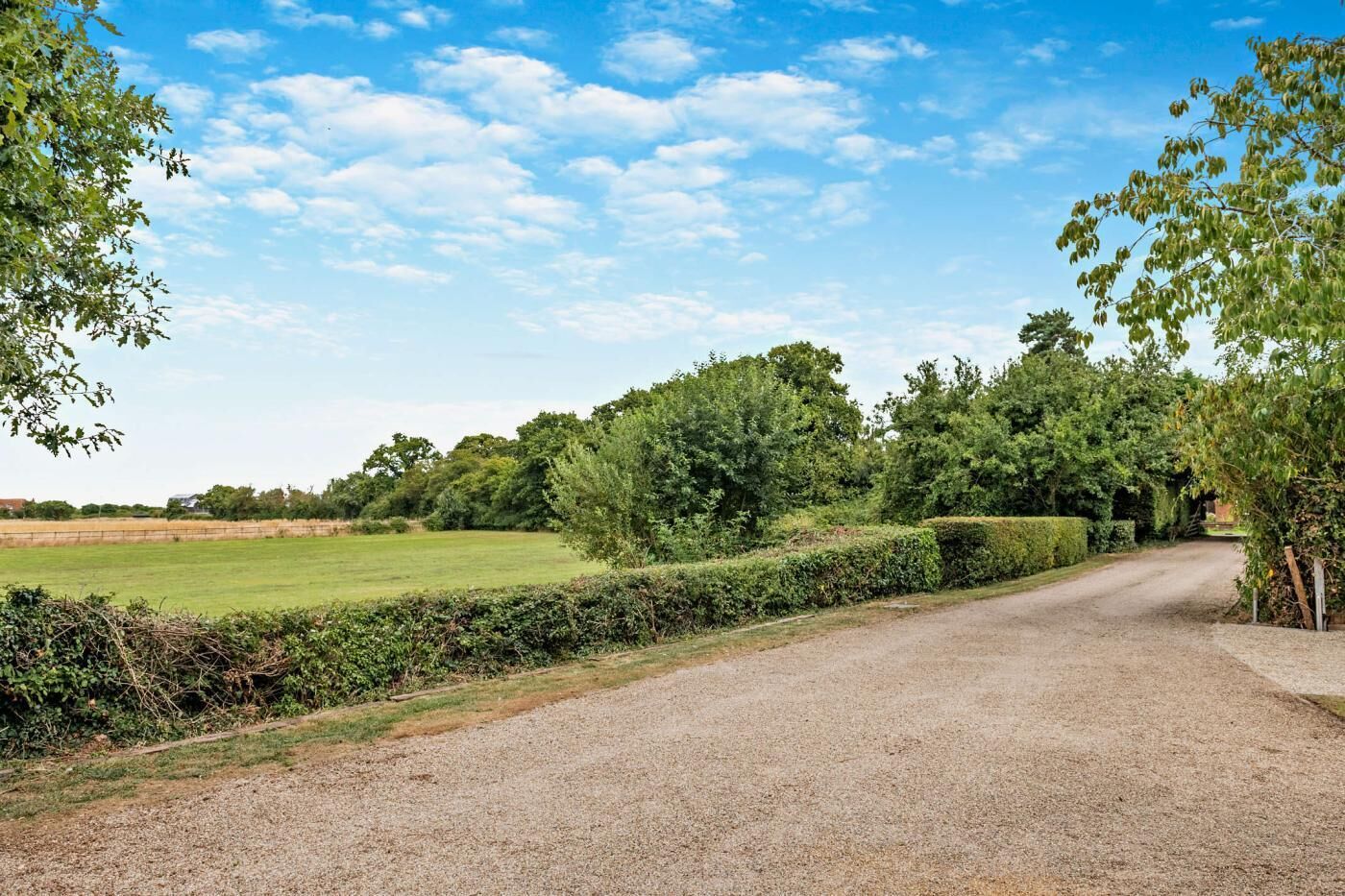 Ein Schotterweg führt zu einem ländlichen Anwesen mit einem von Hecken gesäumten Pfad, Bäumen und blauem Himmel.