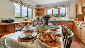 Una cocina moderna con armarios de madera, una mesa de desayuno con croissants, cereal y jugo, y ventanas amplias que ofrecen vistas al exterior.