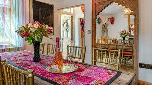Un comedor con una mesa dispuesta para cuatro, con mantel de colores, un florero con flores y una botella. La habitación presenta una decoración tradicional con sillas de madera y un espejo decorativo.