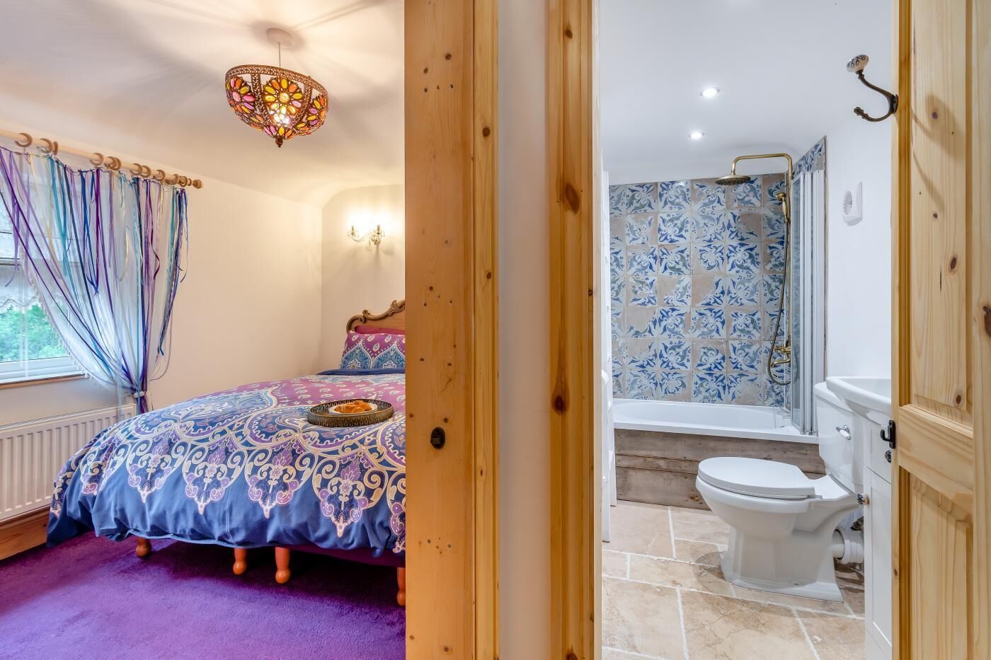 Une chambre à coucher avec un lit, un lustre et une fenêtre avec des rideaux. La pièce est dotée d’une moquette violette et d’une porte en bois qui donne sur une salle de bain équipée d’une baignoire, de toilettes et d’une douche.