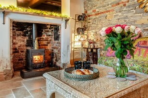A cozy room with a brick fireplace, a decorative mirror, and a table with a floral vase and a plate of croissants. - 4 Bed in Long Bennington (oc-p29872) (Long Bennington)