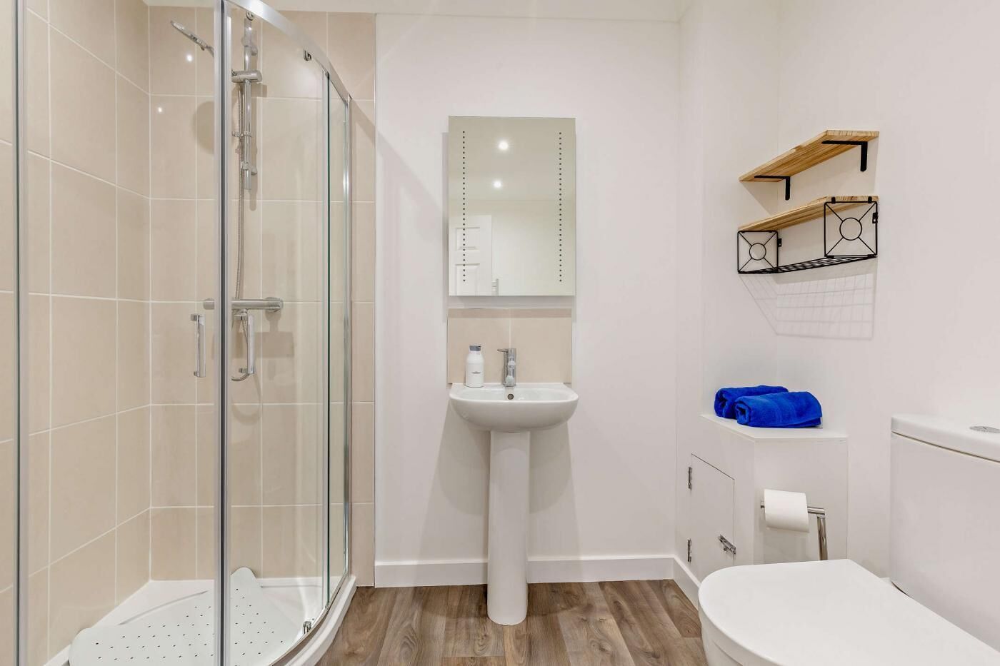 A bathroom with a glass shower enclosure, a pedestal sink, a toilet, and a wooden shelf with towels.