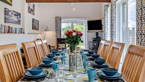 Un coin repas avec une longue table dressée pour six personnes, des chaises en bois, un vase de fleurs et une cheminée en arrière-plan.
