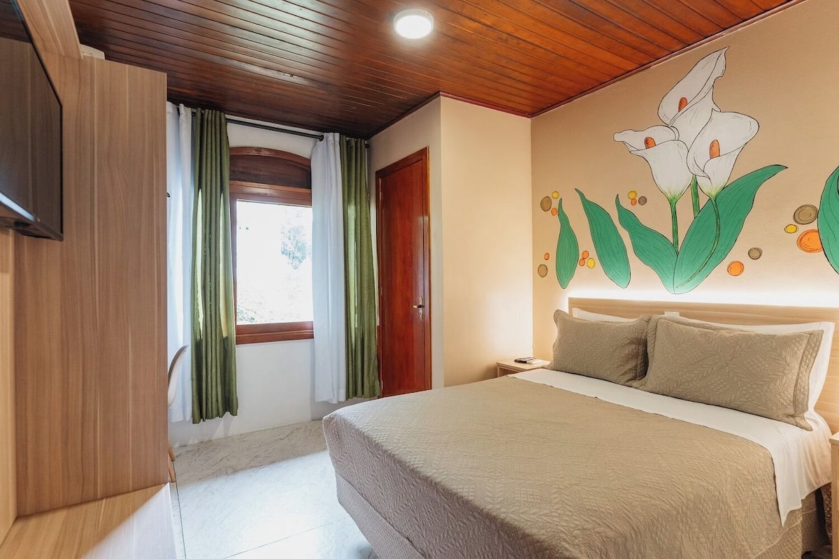 A bedroom with a bed, a wooden ceiling, a wall with a floral mural, and a window with curtains.