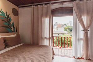 A bedroom with a bed, curtains, and a balcony with a view of buildings and greenery. - POUSADA SANTO ANTÔNIO (Bento Gonçalves)