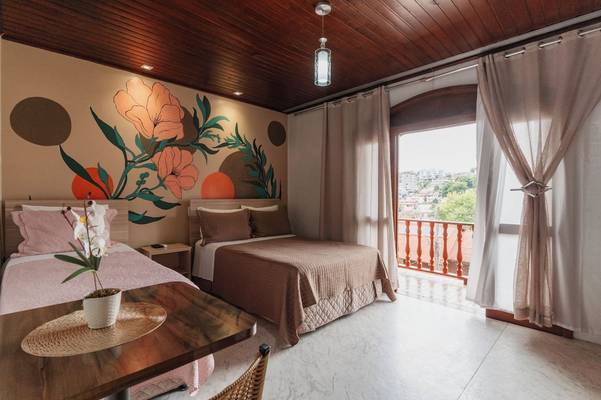A hotel room with a wooden ceiling, a large bed, a smaller bed, a dining table with a plant, and a balcony with curtains.