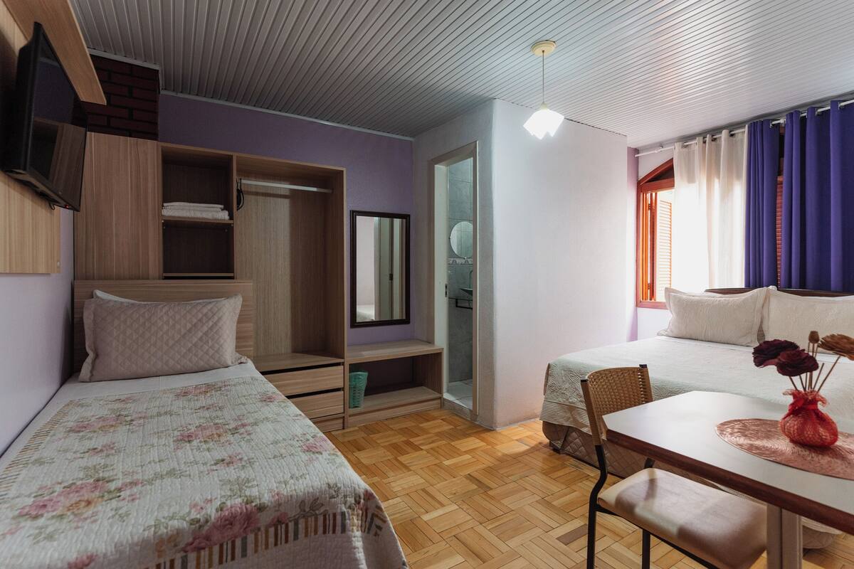 A bedroom with two beds, a wooden wardrobe, a mirror, a table with a vase, and a window with curtains.