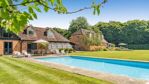 Una piscina con sillas de descanso y un patio, rodeada de césped cuidado y una casa de ladrillo.
