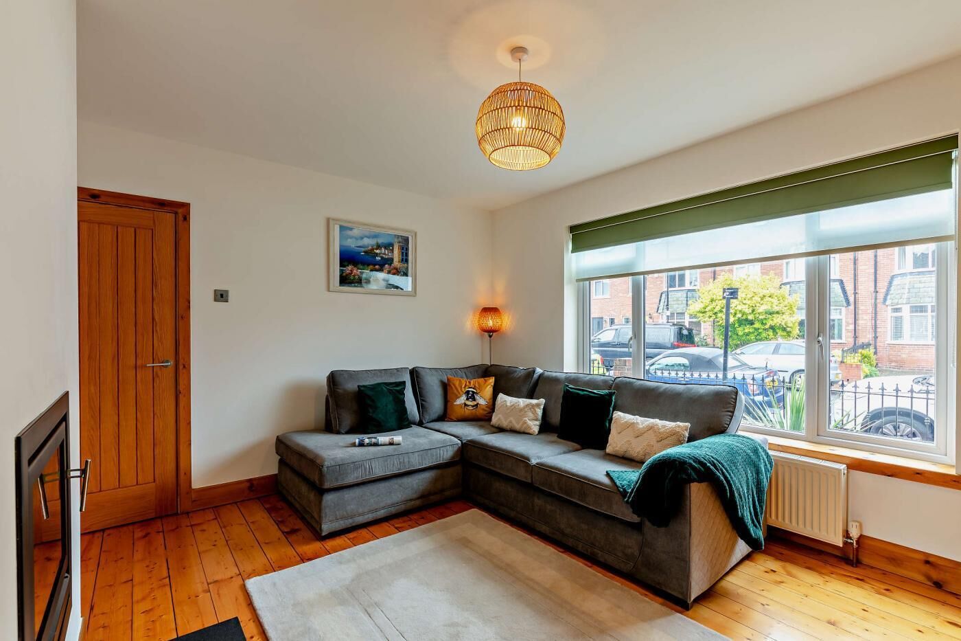 A modern living room with a grey sectional sofa, a wooden floor, a large window with a view of the street, and a hanging light fixture.