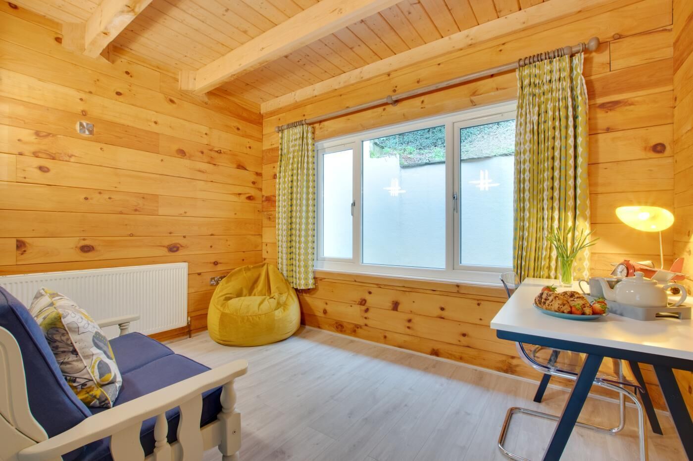 A cozy cabin room with wooden walls, a window with curtains, a sofa, a bean bag, a table with a lamp, and a radiator.