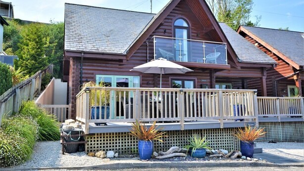 A log cabin with a balcony, a covered patio, and a gravel driveway.