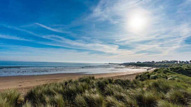 A beach with a clear sky, grassy dunes, and a distant shoreline with buildings. - 4 Bed in Beadnell (oc-s29958) (Beadnell)