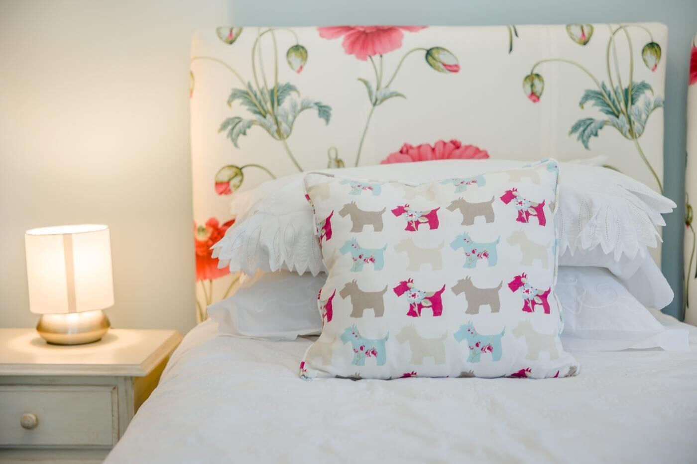 Ein Bett mit weißer Bettwäsche, einem blumigen Kopfteil und einem Zierkissen mit Hunde-Motiven.