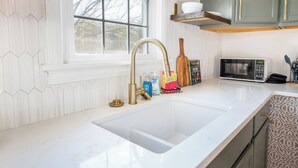 A kitchen with a white countertop, a gold faucet, a microwave, and wooden shelves above the counter. - Sage & Stone cottage-2BR near Downtown Lynchburg & LU (Lynchburg)
