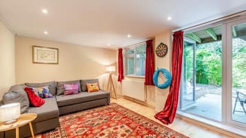 A cozy living room with a grey sofa, colorful cushions, a red patterned rug, and a large window with red curtains.