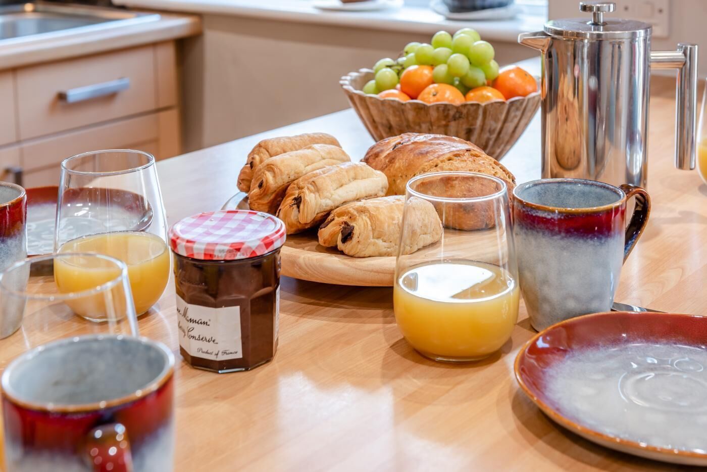 A breakfast setup with croissants, a jar of jam, glasses of orange juice, and a bowl of fruit.