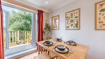 A dining area with a wooden table set for four, a large window with red curtains, and framed botanical prints on the wall.