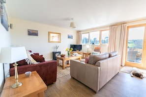 Ein Wohnzimmer mit einer Couch, einem roten Sessel, einem Holztisch, einem Fernseher und einem Fenster mit Vorhängen.