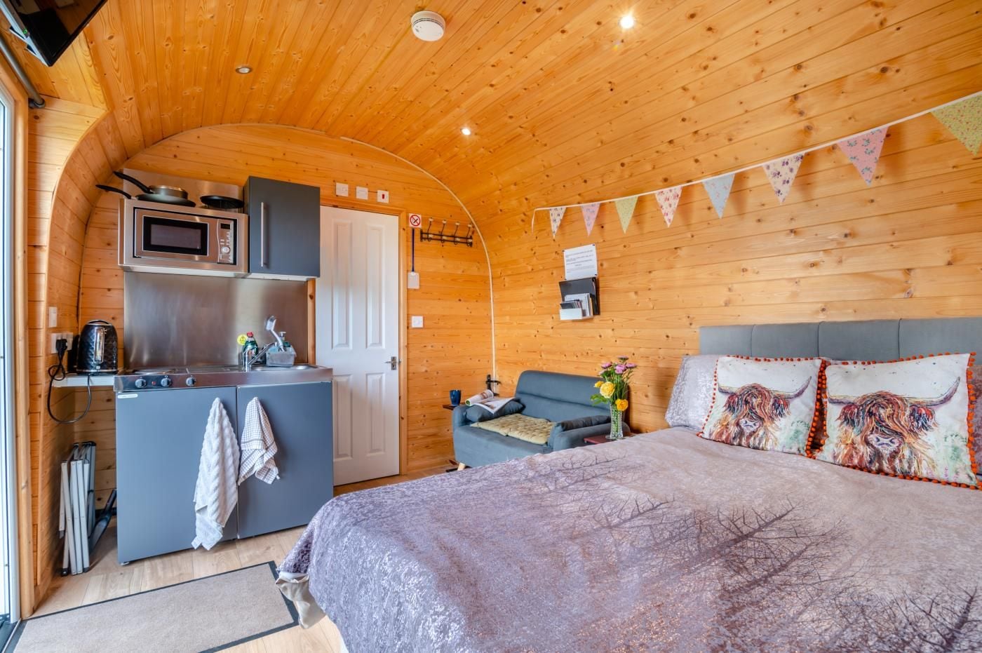 A compact room with a kitchenette, a bed with decorative pillows, and a wooden ceiling.