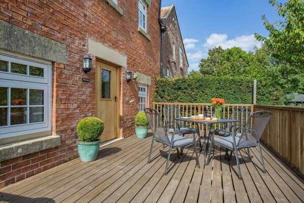 A wooden deck with a table and chairs, a brick building with a door, and potted plants.