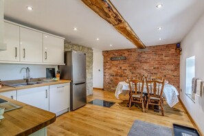 A kitchen with white cabinets, a stainless steel refrigerator, and a dining area with a brick wall and wooden chairs.