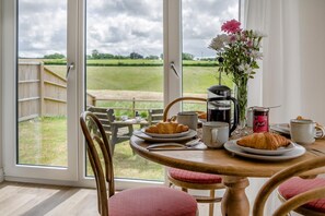 Ein Frühstückstisch mit Croissants, einer Kaffeekanne und Marmelade, mit Blick auf ein Wiesenfeld und einen Holenzaun.