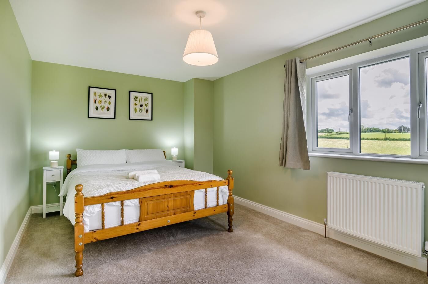Quarto com cama de madeira, paredes verdes, janela com vista para o campo e duas luminárias de cabeceira.