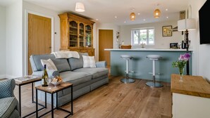 Una sala de estar acogedora con un sofá estampado, un aparador de madera y una zona de cocina con un taburete y un jarrón con flores.