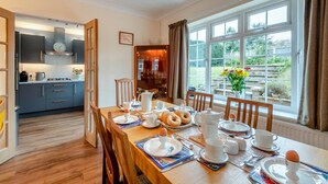 Un comedor con una mesa de madera preparada para el desayuno, una cocina al fondo y vistas a un jardín a través de amplias ventanas.