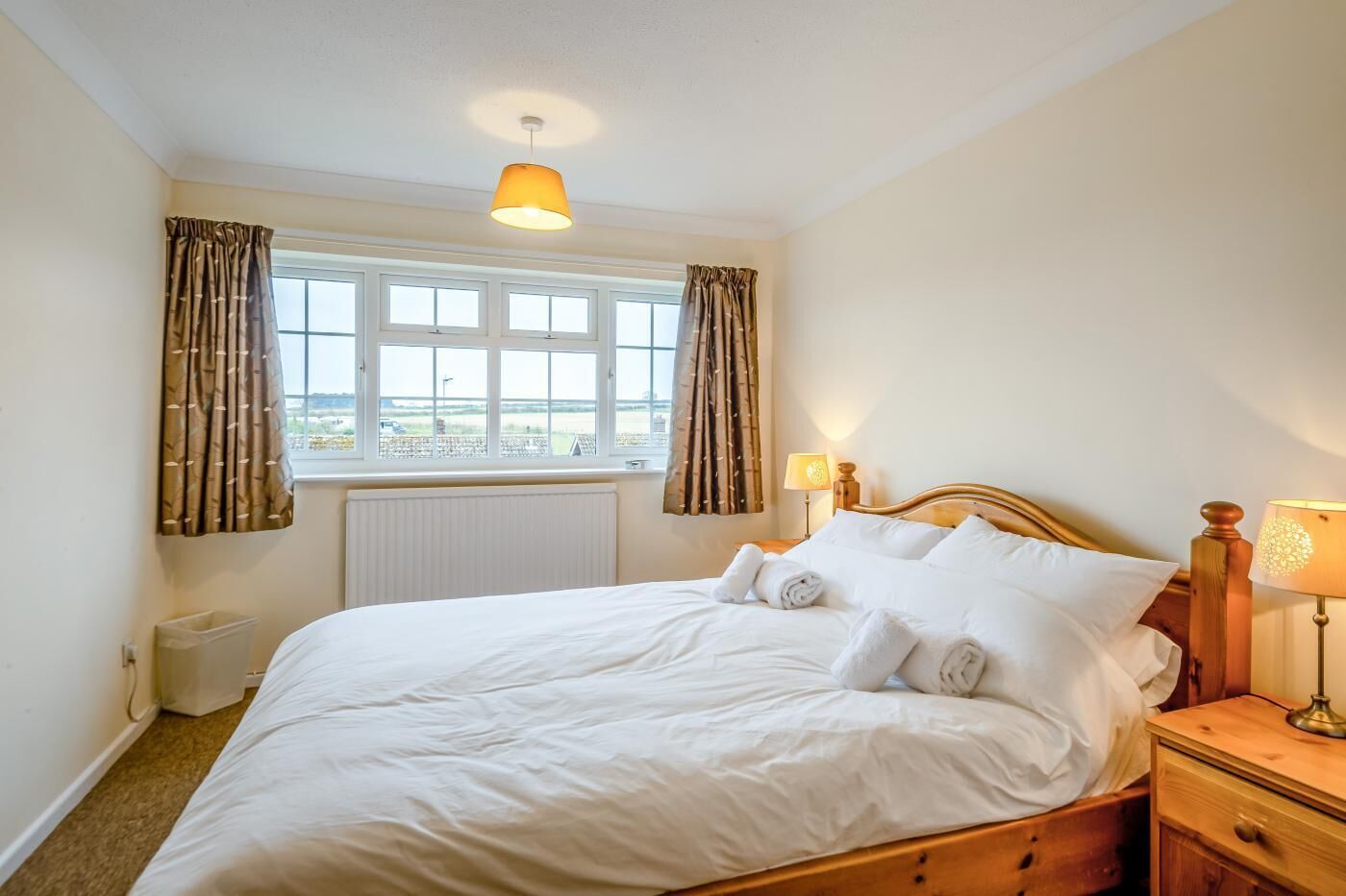 A neatly made bed with white linens and rolled towels, a wooden nightstand with a lamp, and a view of the outside through a window with curtains.