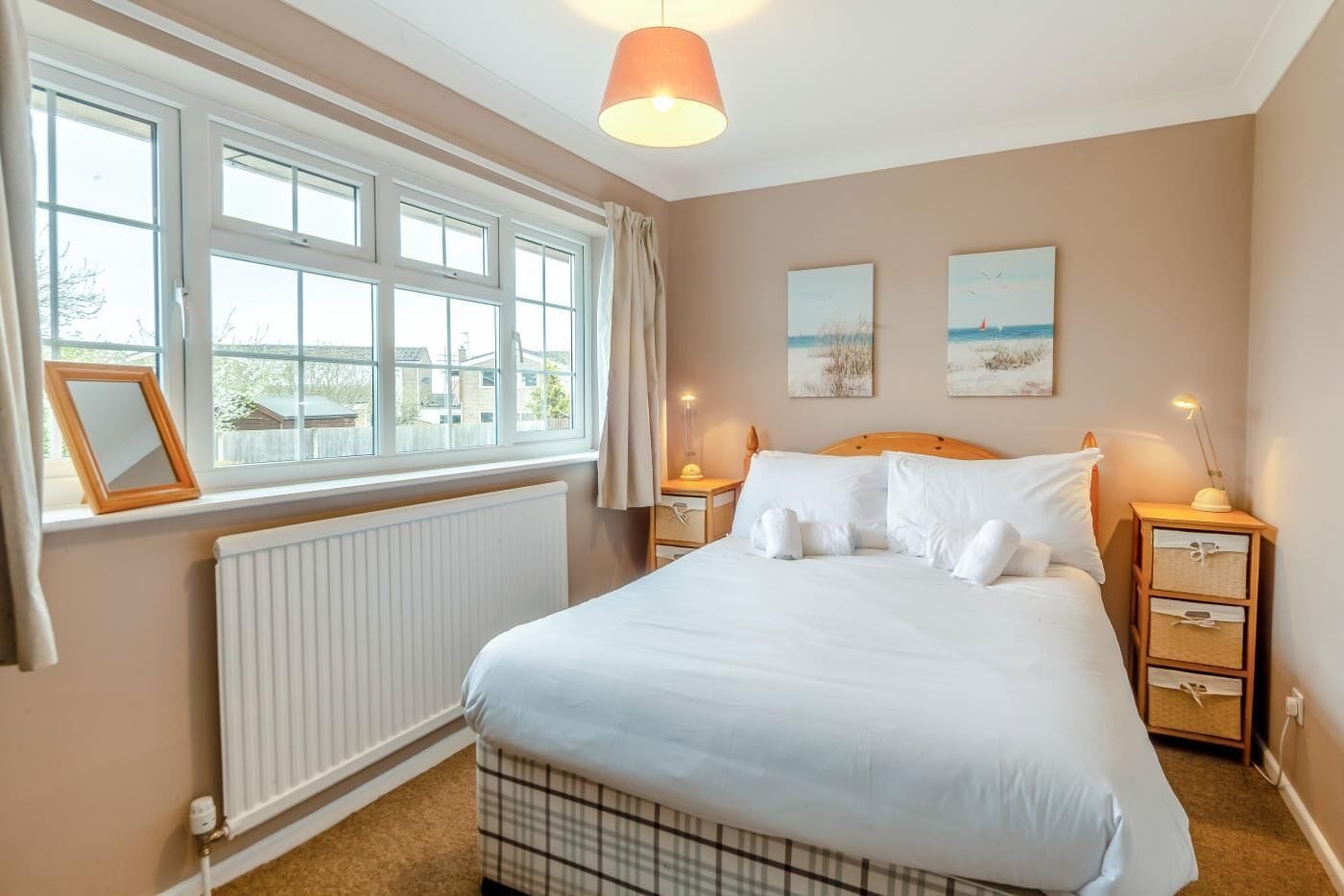 Une chambre avec un grand lit, deux tables de chevet, une fenêtre avec des rideaux, un radiateur et deux tableaux accrochés au mur.