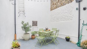 Un pequeño patio exterior con una mesa y sillas, rodeado de macetas con plantas y un muro de ladrillo blanco.