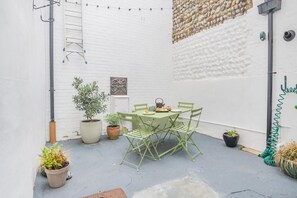 Eine kleine Terrasse im Freien mit Tisch und Stühlen, umgeben von Blumentöpfen und einer weißen Backsteinmauer.